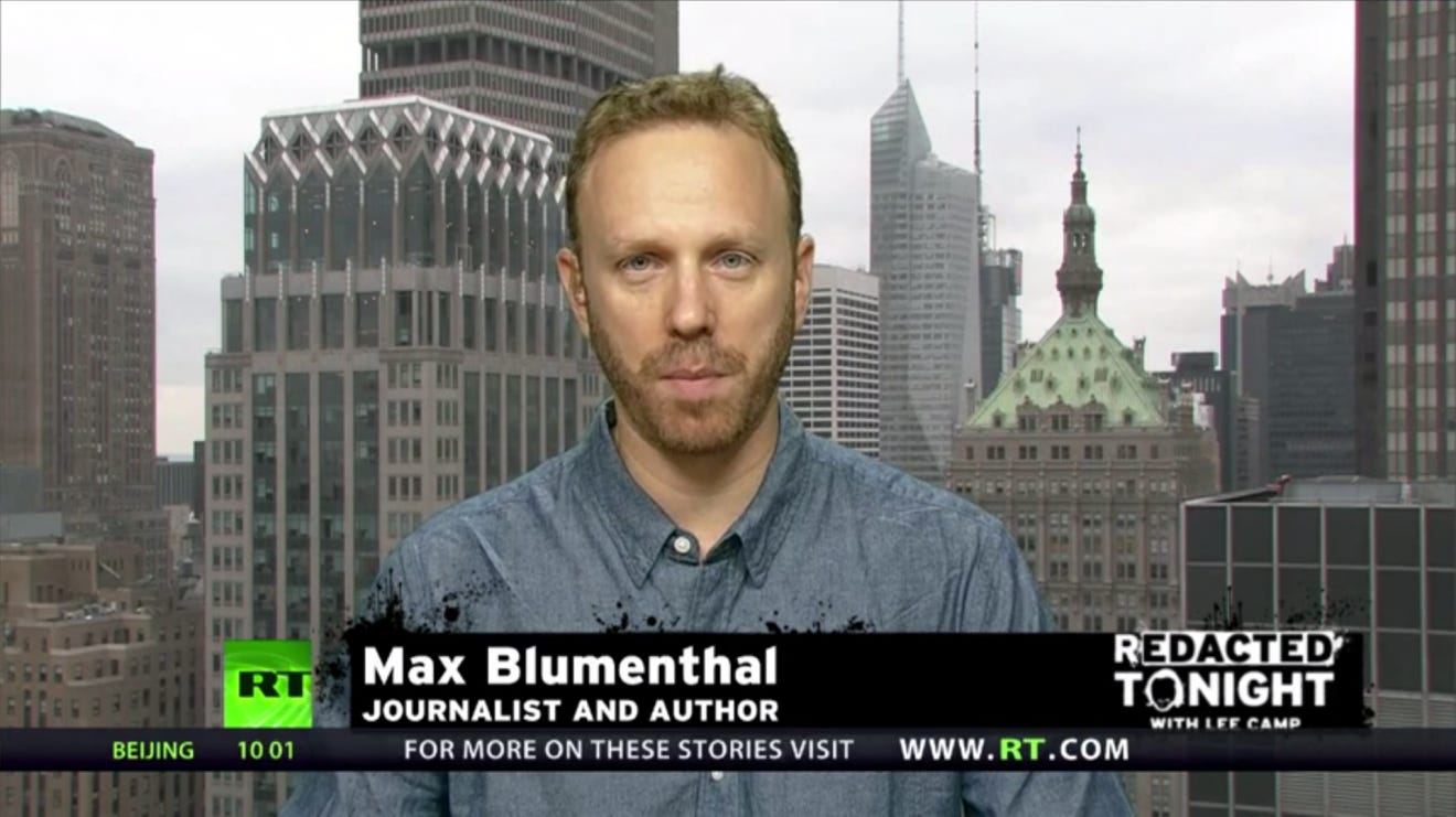 A dead-eyed man with a chyron for RT in front of him and buildings in back.