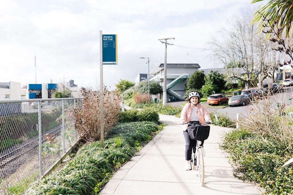 A photograph of Louise on her bike, wearing a pink jacket and a grey helmet, biking along the Western Rail Trail - a separated, landscaped shared path. They look very happy, and it is a beautiful day in Hamilton.