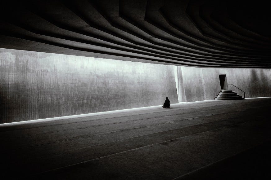 Black and white. A figure in the distance of a concrete empty hall sitting in meditation.
