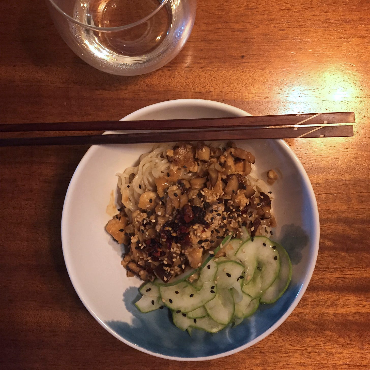 A white and blue bowl of noodles topped with a mix of tofu and mushrooms in a spicy sesame sauce, and thinly sliced quick pickles. Resting on the edge of the bowl is a set of dark wooden chopsticks, and a stemless glass of white wine is just visible and the top of the frame.
