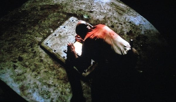 John Locke (Terry O’Quinn), his shirt stained with blood, peers into the hatch. There is a light within.