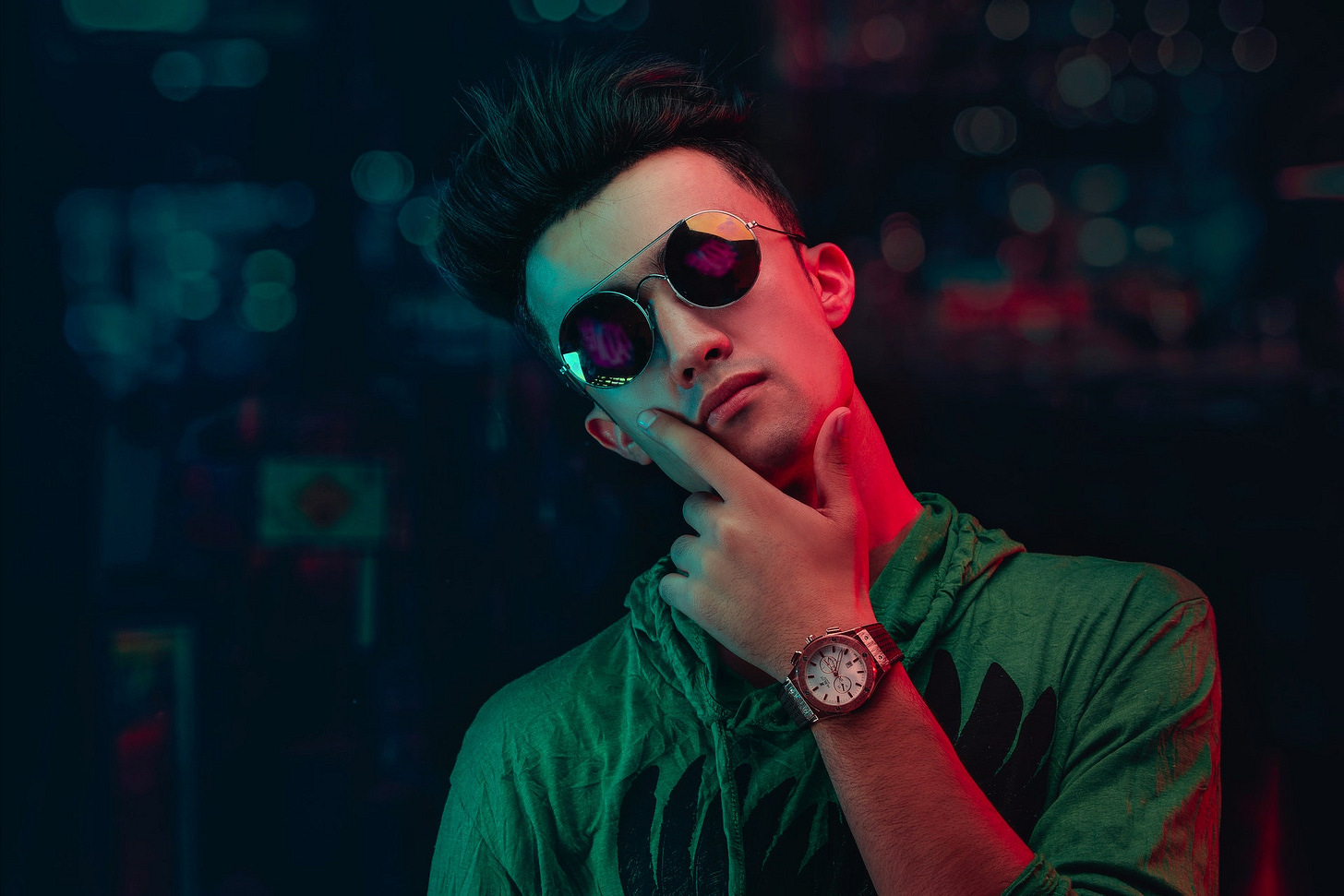 A man in a green shirt wearing a watch and reflective sunglasses with his hand on his chin wondering