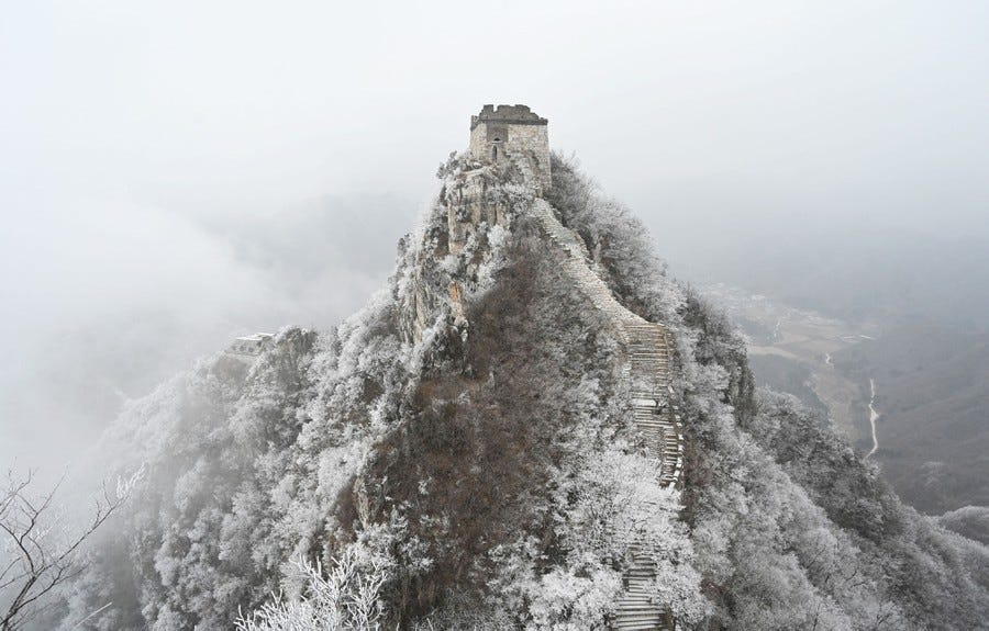 A stone tower and steps on a wide stone wall ascend to a steep peak covered in frost and snow.