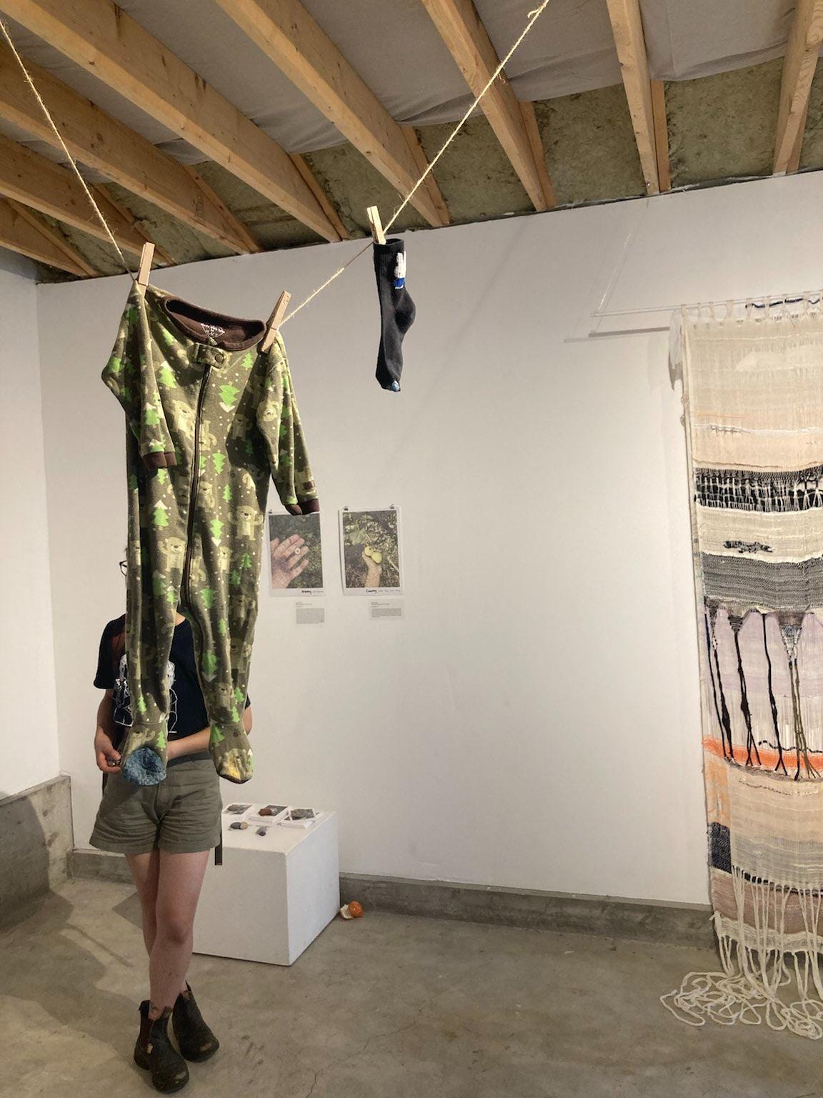 An image of Khanh's pieces at the exhibit. A forest green baby's onesie with 2 mending repairs on the toes are on a clothes line, accompanied by a black sock with Miffy the bunny on it.