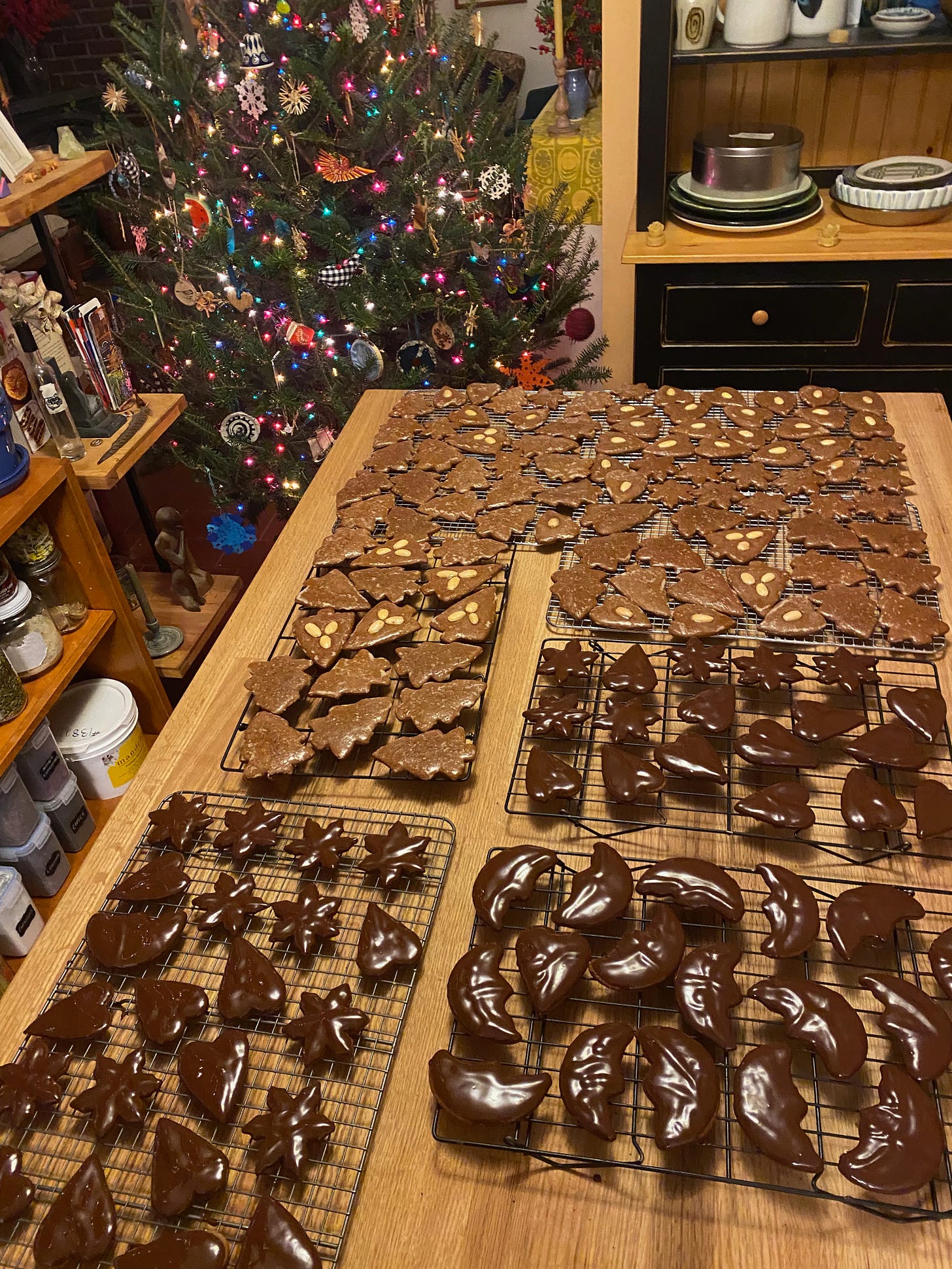 150+ gingerbread cookies, some dipped in chocolate, spread out on cooling racks on a kitchen island. A decorated Christams tree is visible in the background.