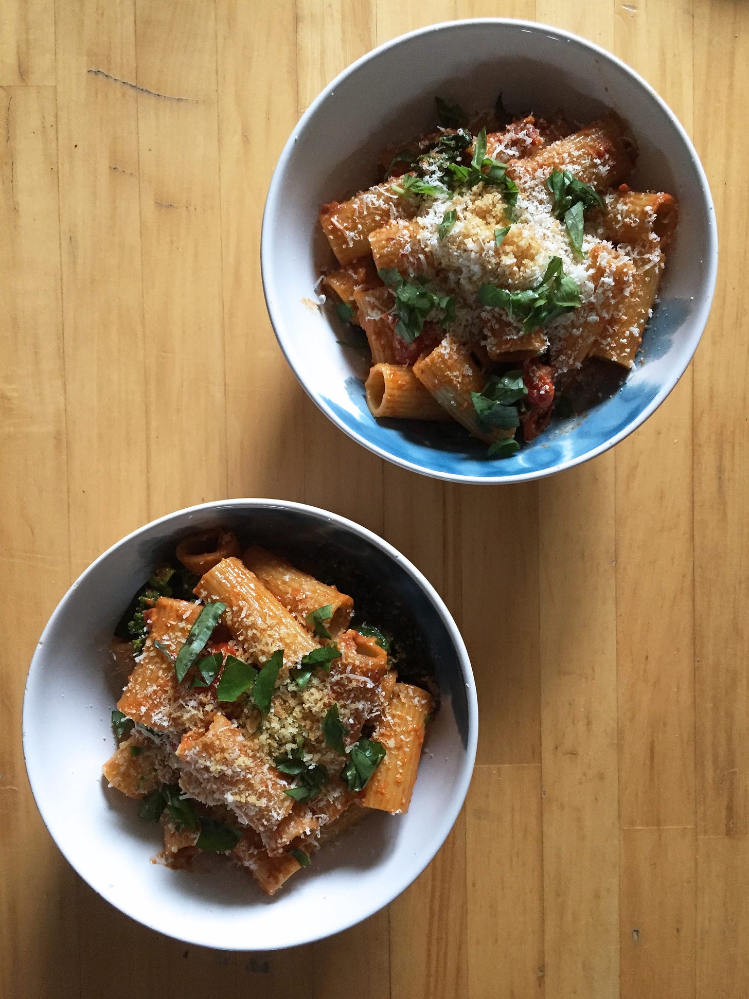 two white bowls of rigatoni in a dark red pesto sauce, with shreds of basil, parmesan, and toasted panko crumbs on top.