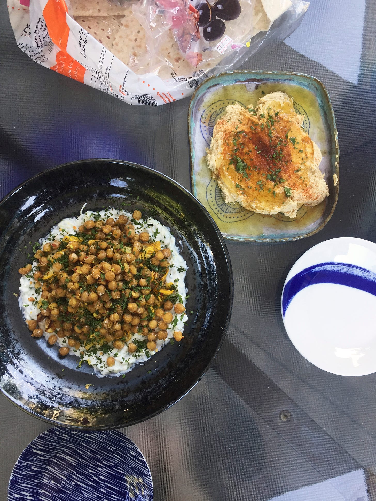 A shallow black dish full of the tzatziki, zucchini, and chickpeas, with herbs and lemon zest on the top. Another smaller, shallow blue dish has a smear of hummus in the centre, coated in olive oil and spices and chopped herbs. At the top of the frame is a bag of taftoon, and two small plates are at the lower edge of the frame.