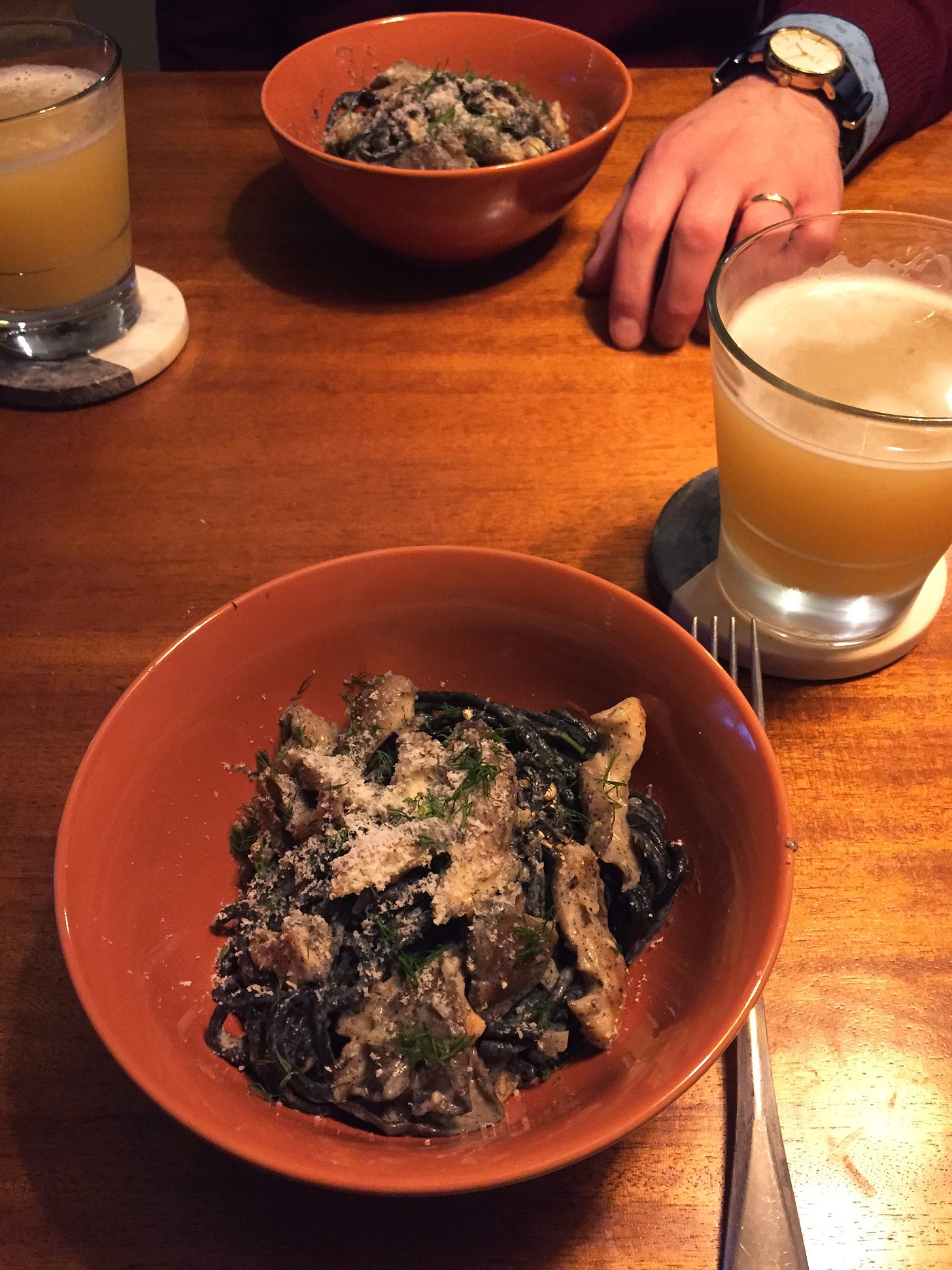 Two orange bowls across from each other, each filled with black noodles and large pieces of oyster mushroom in a light cream sauce. Parmesan and fresh dill dust the top, and two glasses of IPA are on coasters next to the bowls.