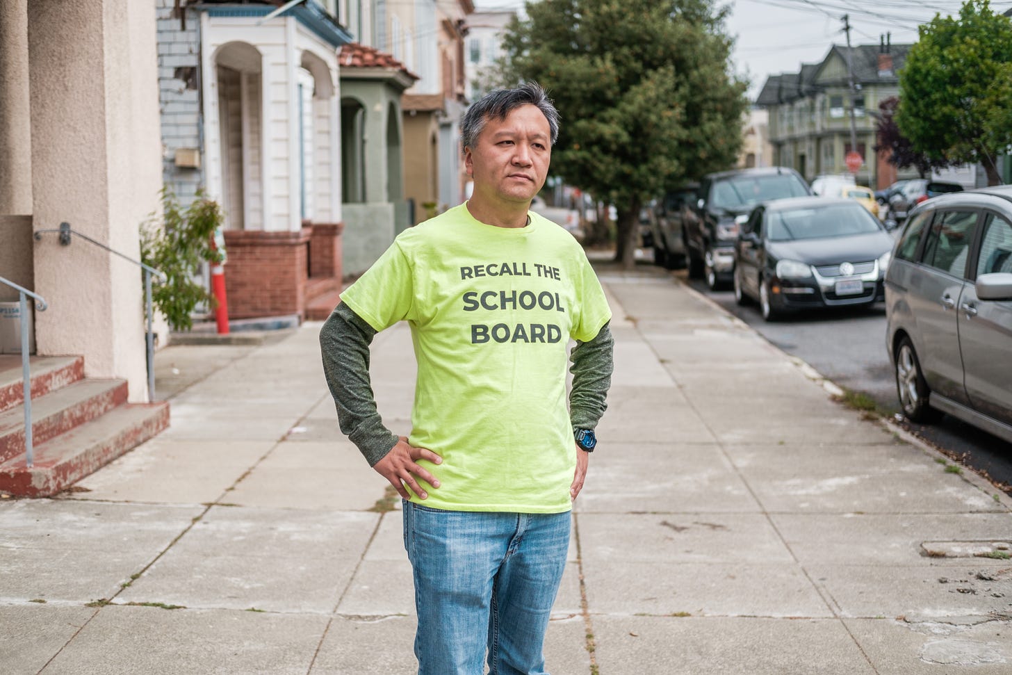 Kit Lam, a Chinese man, stands on a sidewalk with his hands at his hips, wearing a shirt that says "RECALL THE SCHOOL BOARD"