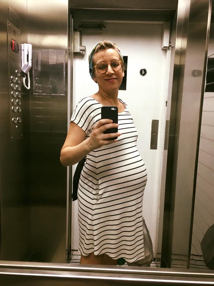 foto de uma mulher, eu no caso, com uma barriga de gravidez de tamanho razoável, usando um vestido branco listrado de preto, uma faixa colorida nos cabelos, fazendo uma selfie de elevador sorrindo e com os olhos baixos