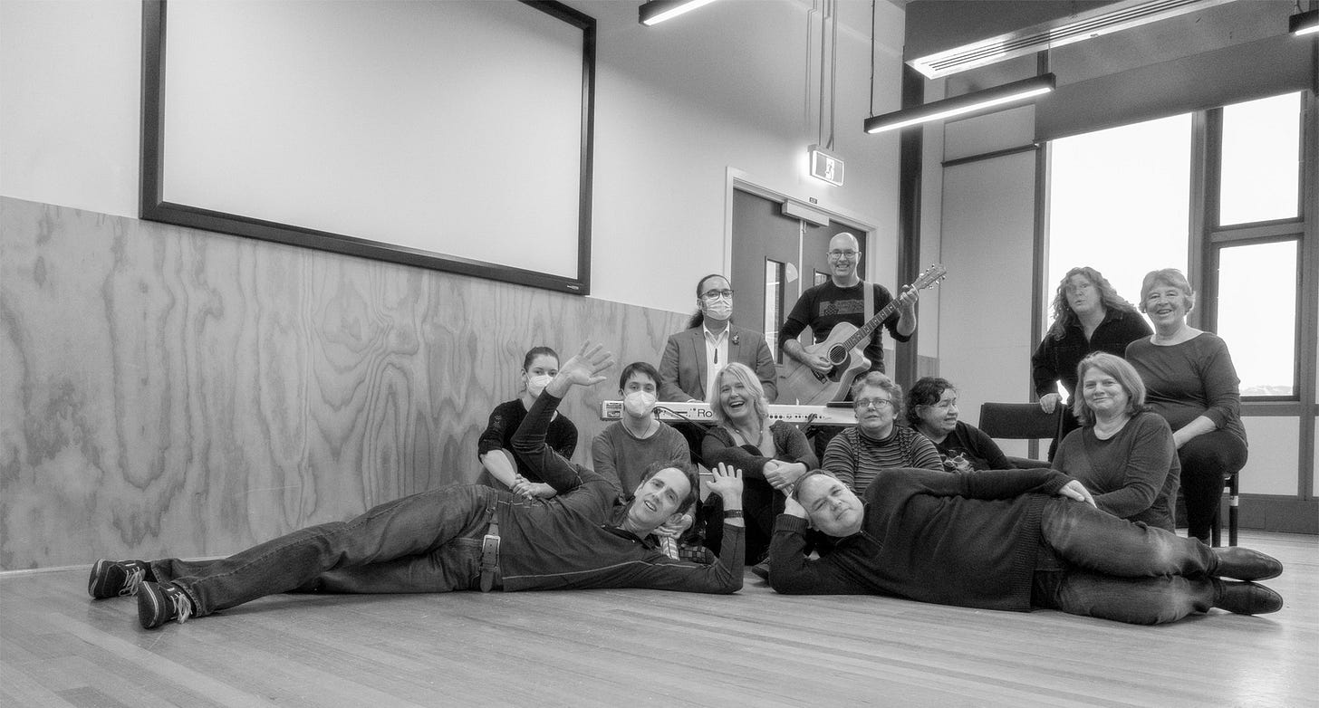 Black and white picture of improvisors in a workshop surrounding a piano, mostly sitting or lying down, smiling at the camera