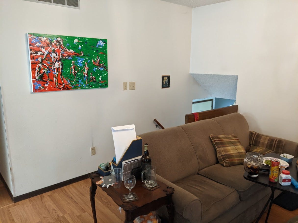 A living room couch. On small tables adjacent to it there are empty cups, a plate and small bowl with edamame husks, bottles and cans, medicine bottles. On the wall behind the couch, a vivid red and green painting, and a photograph too small to see.
