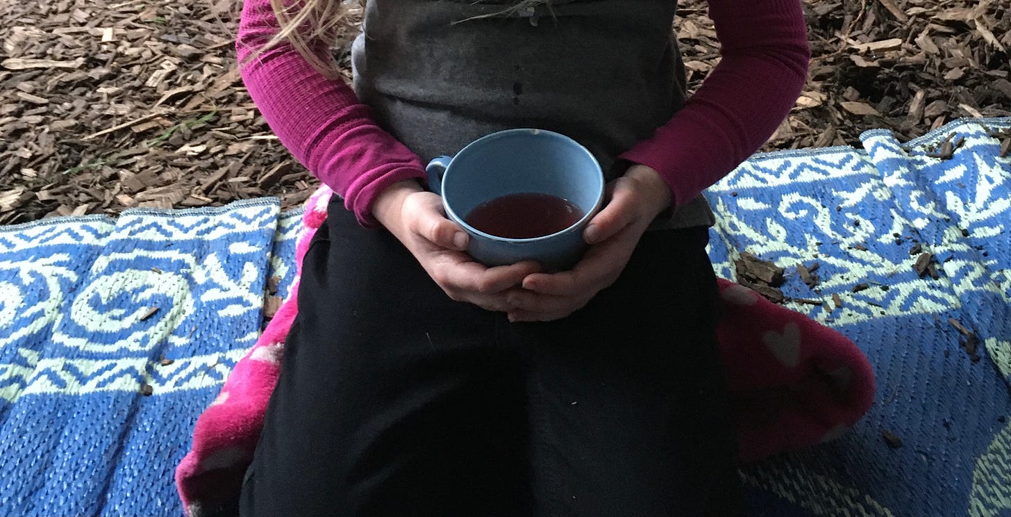 cropped photo of child sitting with their legs tucked under them, wearing black pants and a gray shirt with pink long sleeves, light-colored hands holding a blue porcelain cup with some pink tea in it, gently holding the cup, child is sitting on top of blue and patterned plastic mat and behind child and mat are wood chips. child is also sitting on top of fuzzy pink jacket. 