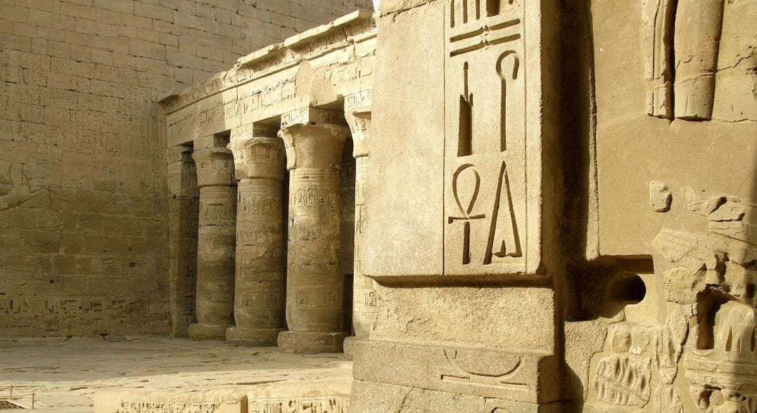 Four large ancient Egyptian hieroglyphs are engraved on the wall of a temple with columns in the background.