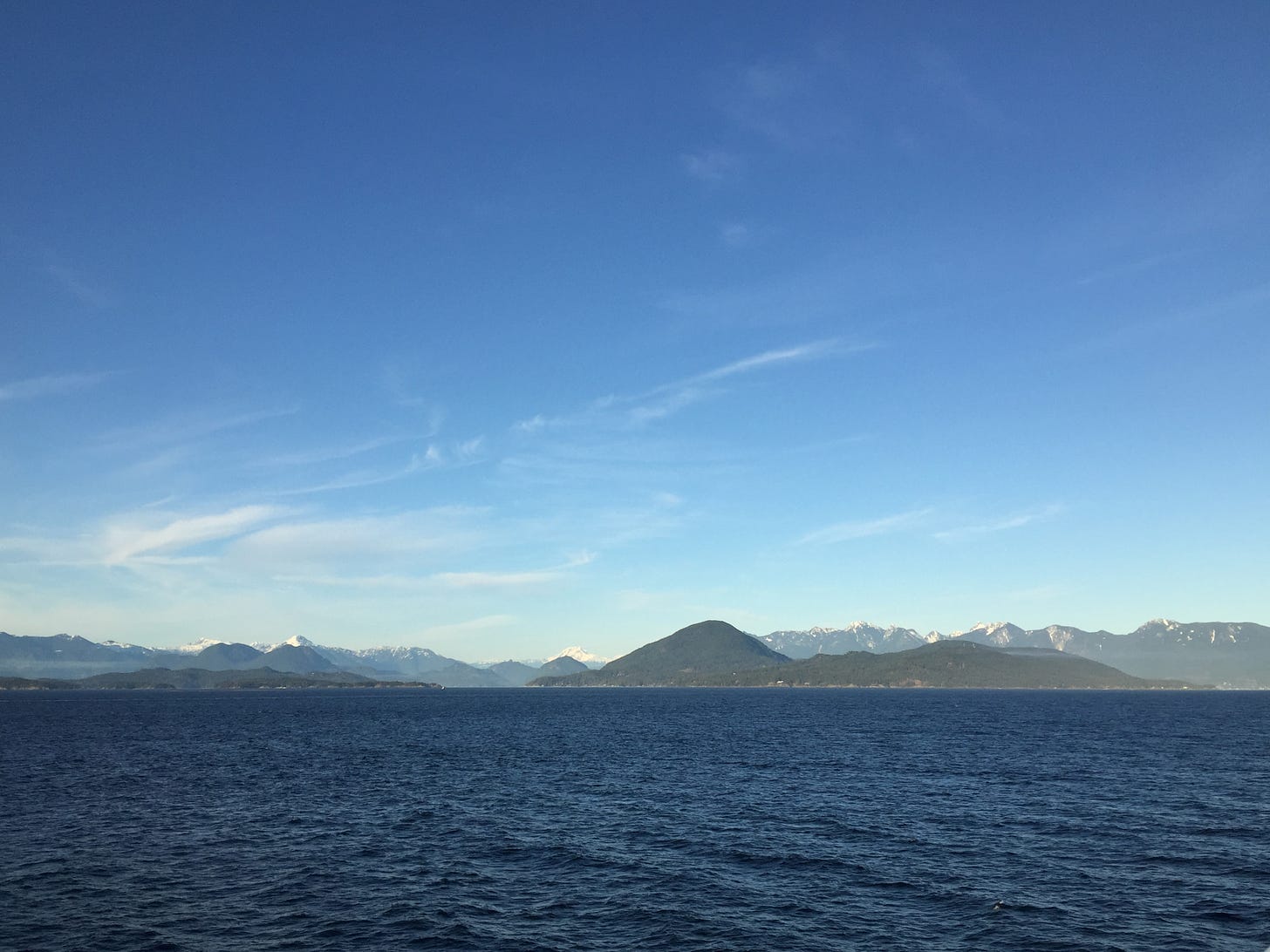 A view of the ocean and mountains taken from the ferry. The sky is blue with wisps of cloud and the ocean is dark blue with small waves.