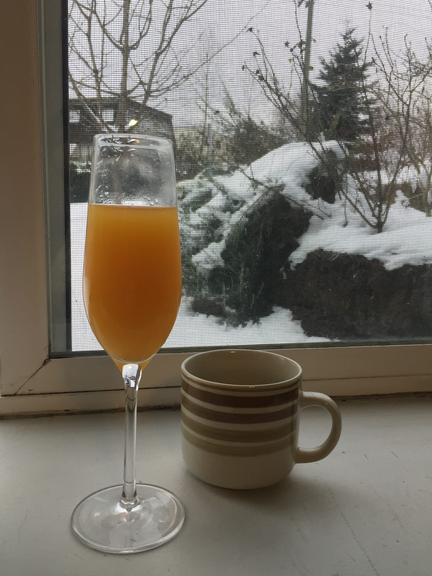 in front of a window showing a snowy yard, a mimosa in a champagne flute and a cup of coffee in a striped brown and beige mug sit on the windowsill.