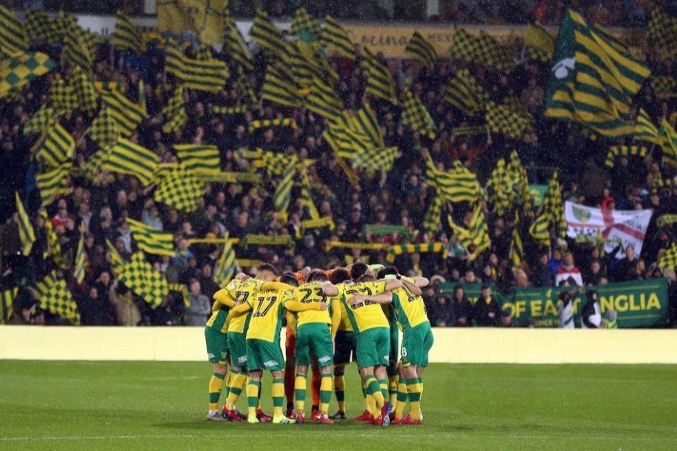 An image of Norwich City players in yellow and green huddled in front of a crowd of spectators waving banners and flags