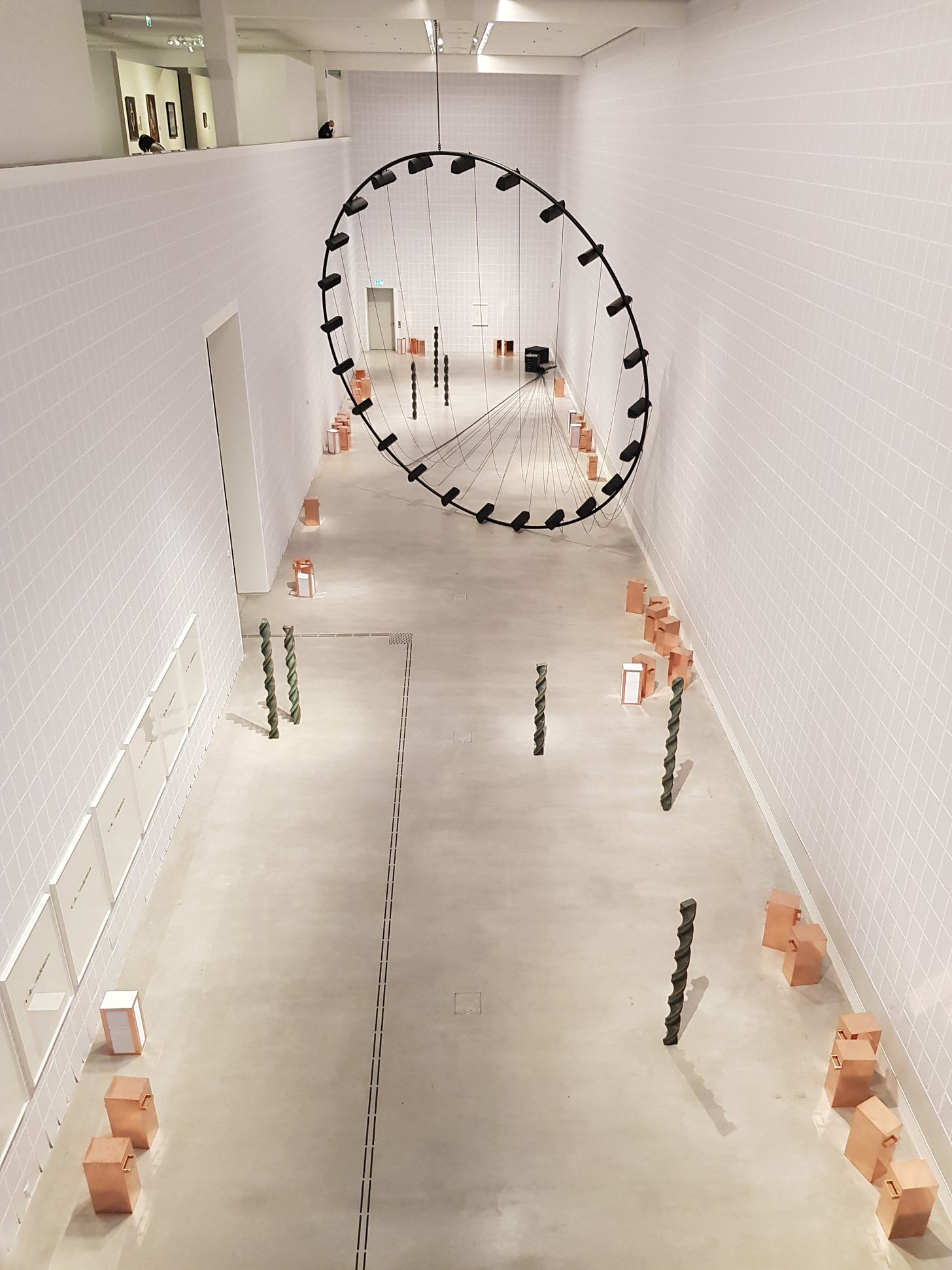 A view of an art exhibition, with a large black steel circle in the middle of the hall and other small objects around the room. The perspective is from the second floor looking down and across the space.