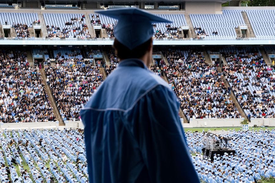 A back view of a person wearing a graduation cap and gown. In the background, rows of graduates sit on a football stadium field.