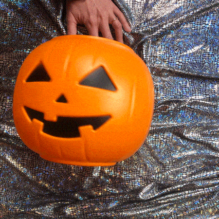 An animated photo loop of an orange plastic blow mold Halloween bucket in the shape of a jack-o-lantern on top of shiny silver fabric with a white-skinned hand ever so slightly moving it back and forth and back and forth. The hand and the fabric are in focus while the pumpkin is blurred. 