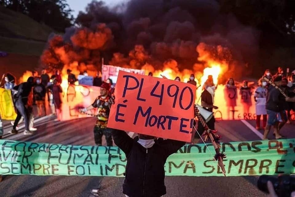 A protest in a street with a person holding up a sign that says 'PL490 E Morte' in front of a banner and a crowd of people in front of smoke grenades that have gone off