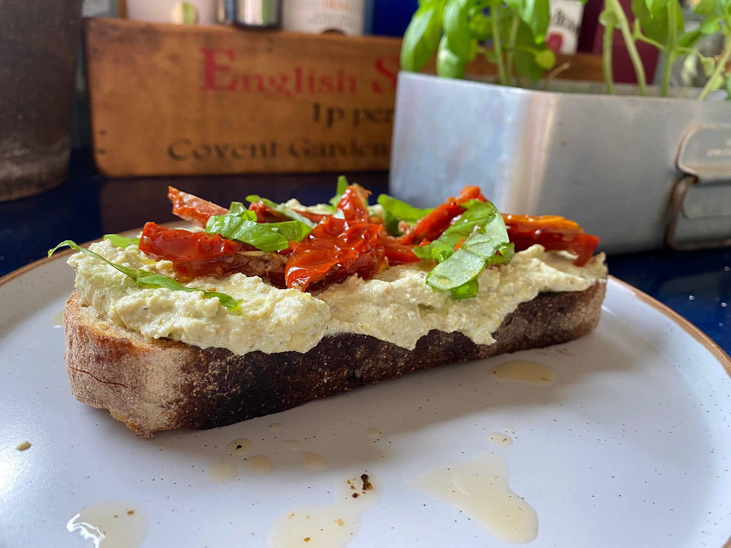 Slice of toast topped with cream cheese and sliced red sun-dried tomatoes and torn basil leaves. A silver container with basil plants is placed behind the plate on the worktop.