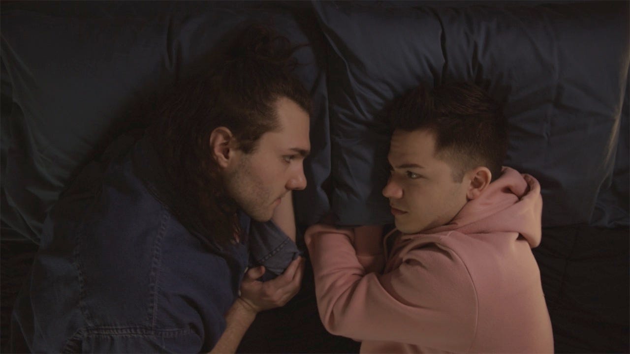 Two young men wearing sweatshirts lie on a bed, intimately talking and gazing at each other. The actors Benjamin Mills and Derek Quesada portray reimagined characters in love based on the characters of Benwick and Louisa from Jane Austen's 'Persuasion.' 
