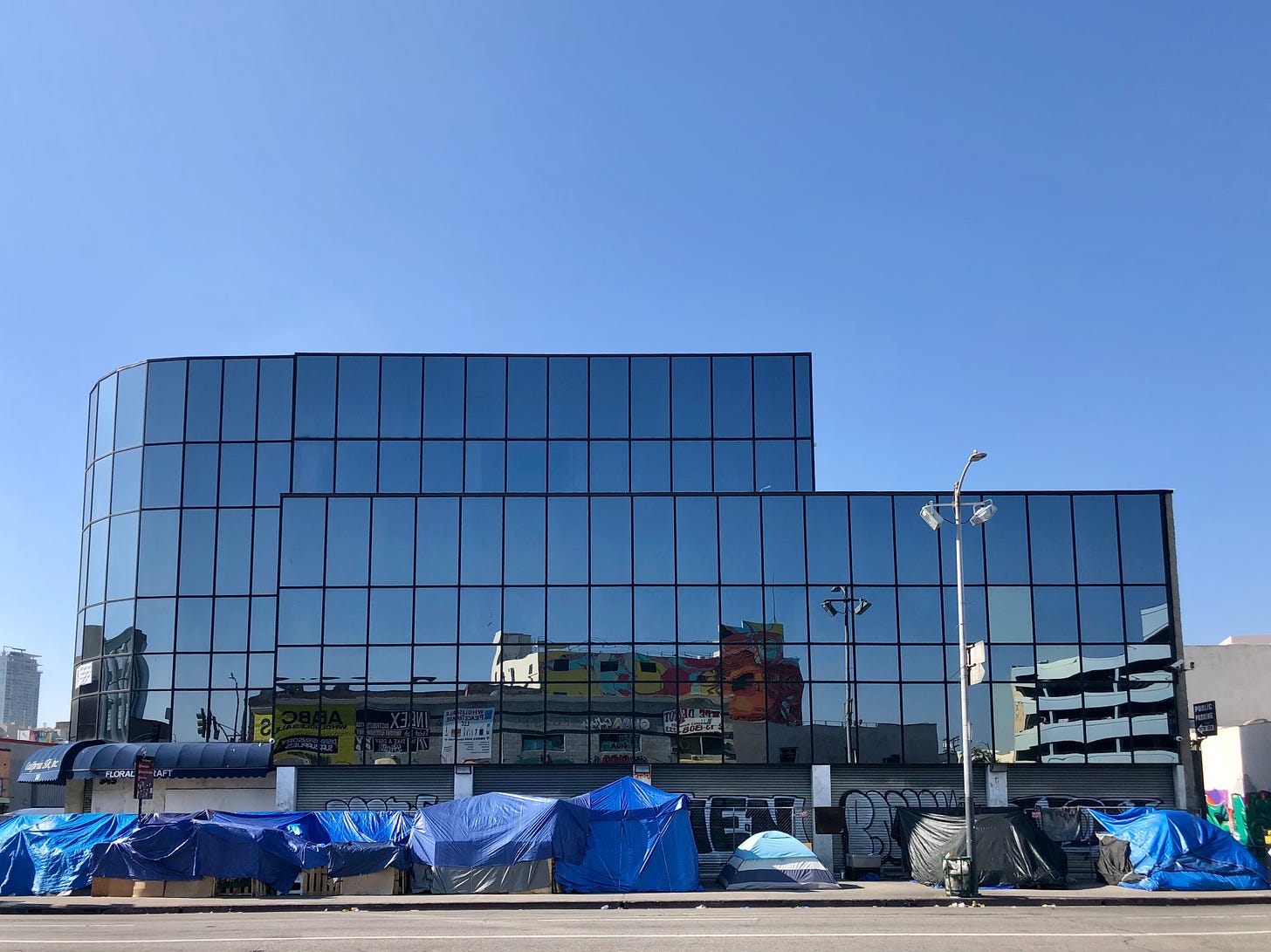 A three story, reflective glass building in Skid Row, Los Angeles surronded by makeshift shelters draped in blue tarps.