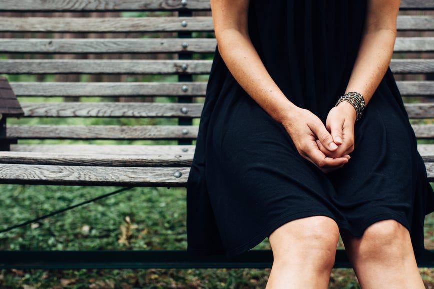 the lower half of a woman in a black dress sitting on a park bench, alone, with her hands in her lap