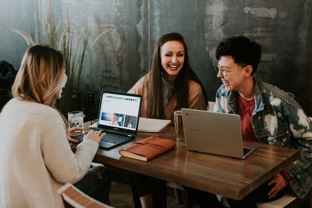 three people sitting around a table with laptops, laughing