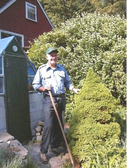white man in blue shirt and green hat standing in garden with tool
