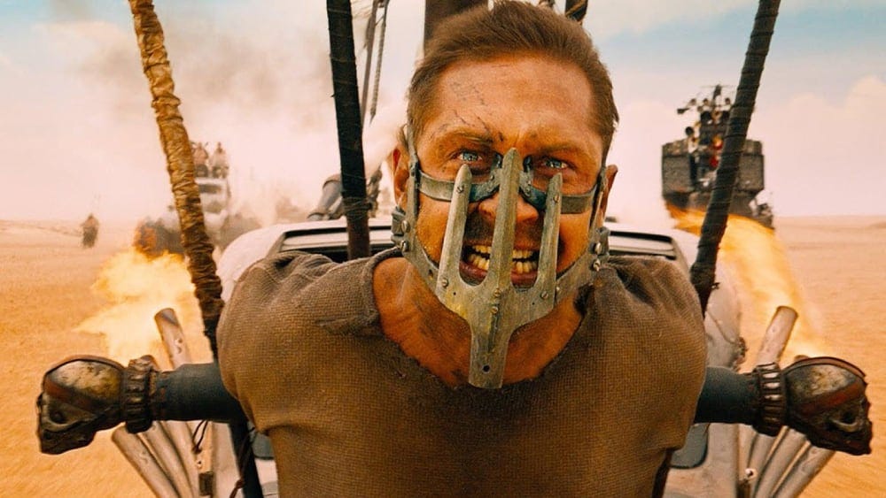 Tom Hardy in a mask, strapped to the front of a car, in Mad Max: Fury Road
