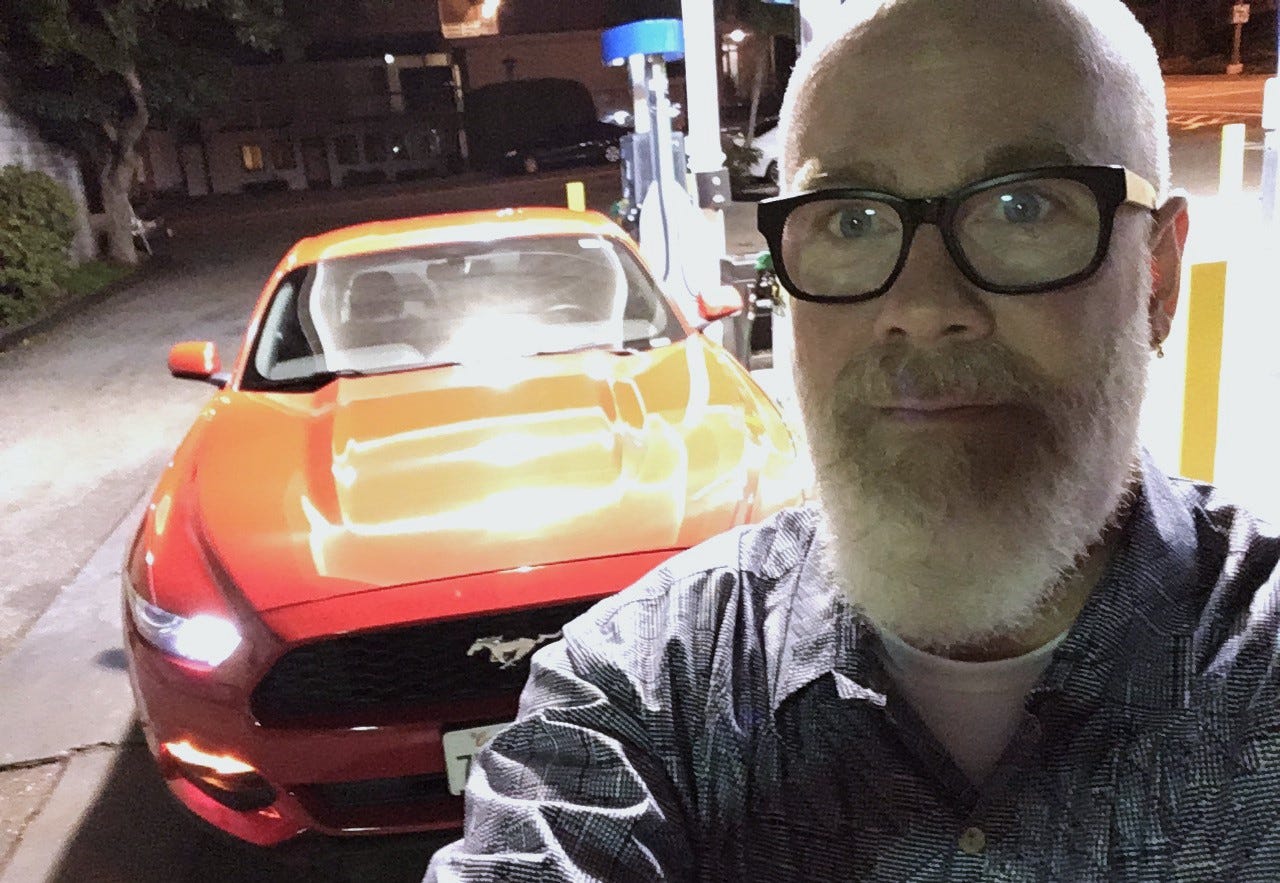 On a darkened commercial street, a gray-bearded man with glasses takes a selfie with gas pumps and a bright red Ford Mustang behind him.
