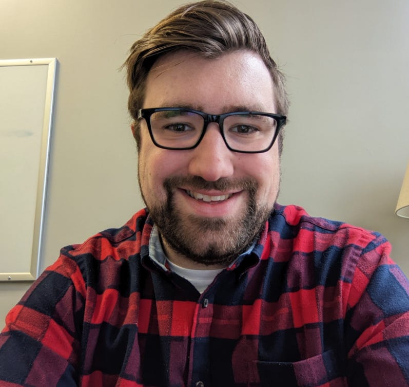Picture of Joe Ray with brown hair and beard, brown glasses, and a red plaid flannel shirt. Sitting in an office with light-gray walls.
