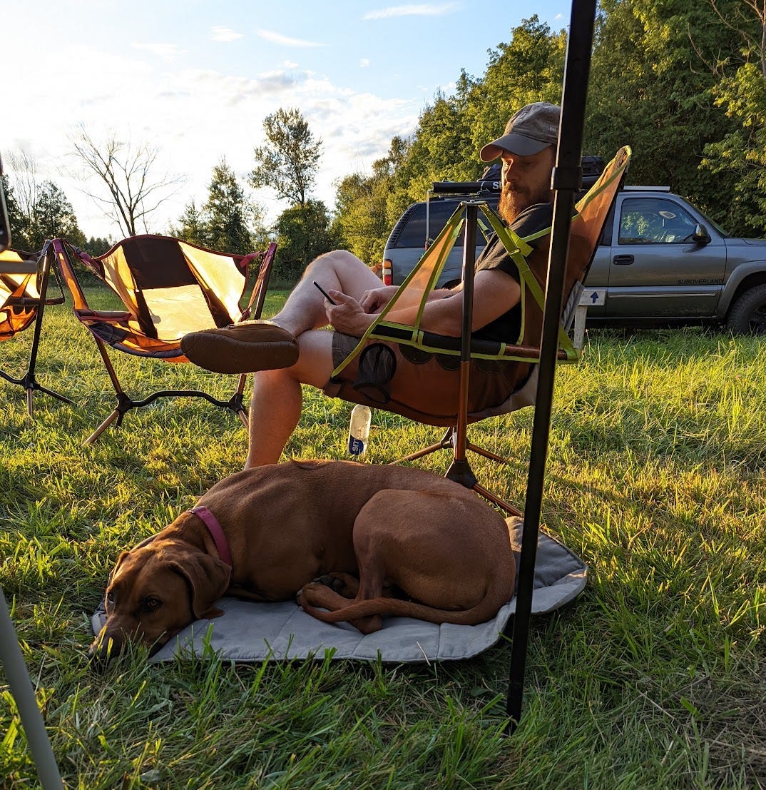 A picture of Rusty in a camp chair, at the golden hour in a beautiful green field looking down at Sammy, the most handsome Ridgeback, sleeping next to me. I look very beardy and relaxed.