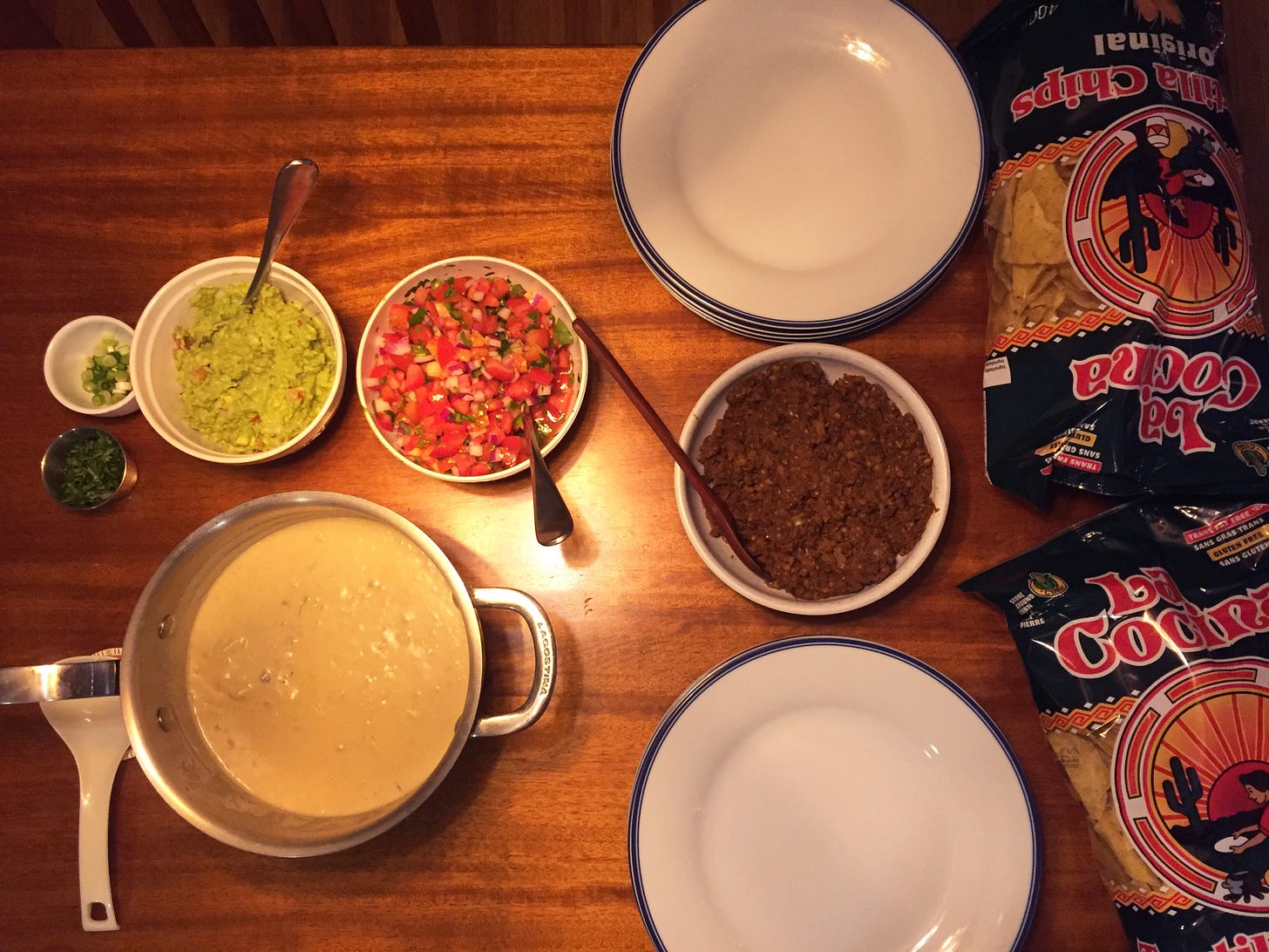 From above, a table full of nacho fixings: bags of chips on the right, two stacks of plates with a bowl of lentil meat in between them, a pot of queso with a spoon beside it, and above the pot, bowls with pico de gallo, guacamole, chopped cilantro, and green onions.