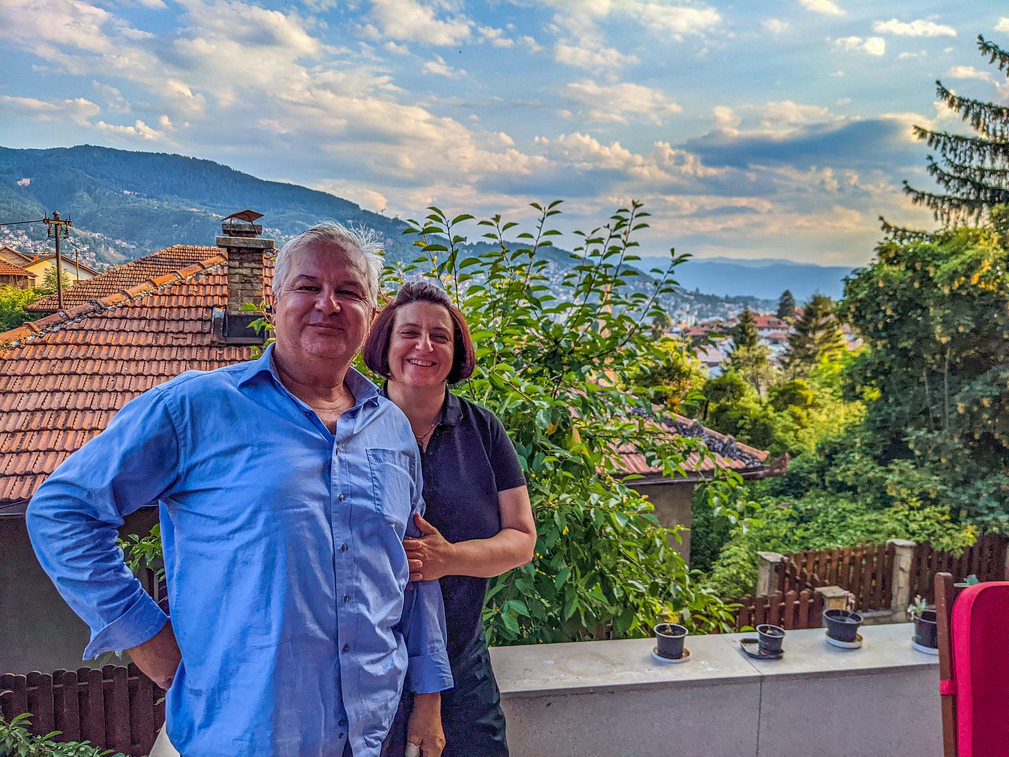 Tarik and Edina standing on their porch, a tree-filled view of Sarajevo behind them. 