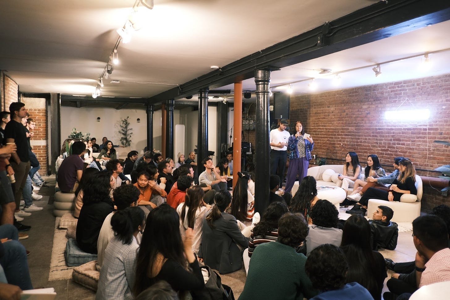 A crowd is gathered in an open room. Most people are sitting on cushions in the floor and some people are standing against the back wall. Six people are facing the audience at the front of the room. One person is speaking into a microphone.