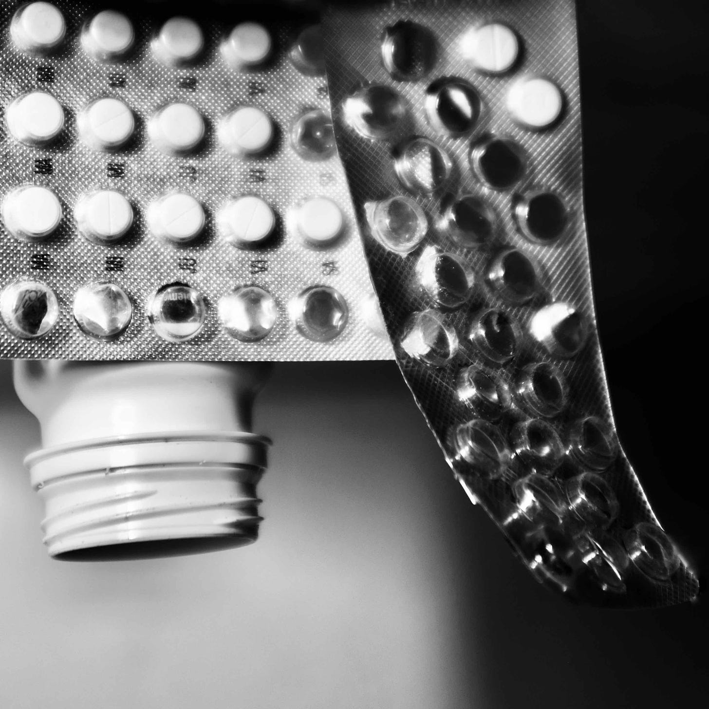 A black & white photograph of an upside-down pill bottle facing down from the left-hand side. Above it are two overlapping half-empty sheets of pills. 