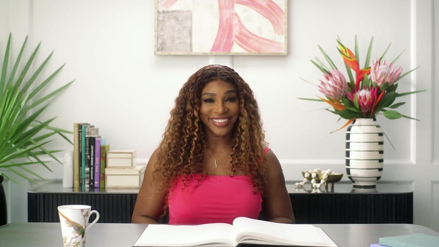 Serena Williams sitting at a desk during a Vogue interview. She is wearing either a pink top or pink dress. Her hair is down and she is smiling.