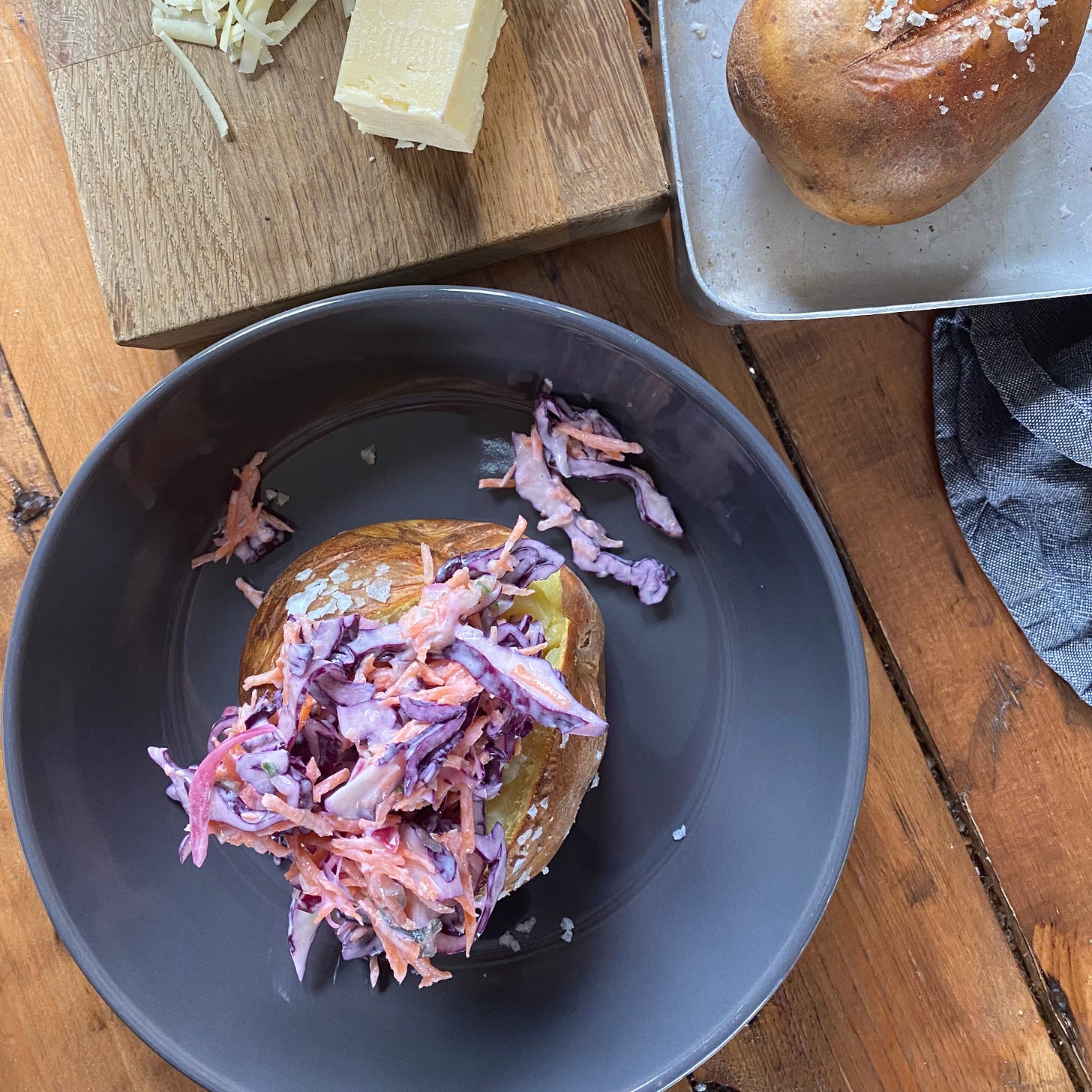 Jacket potato topped with cheese and coleslaw, another potato in a baking tray to the side. 