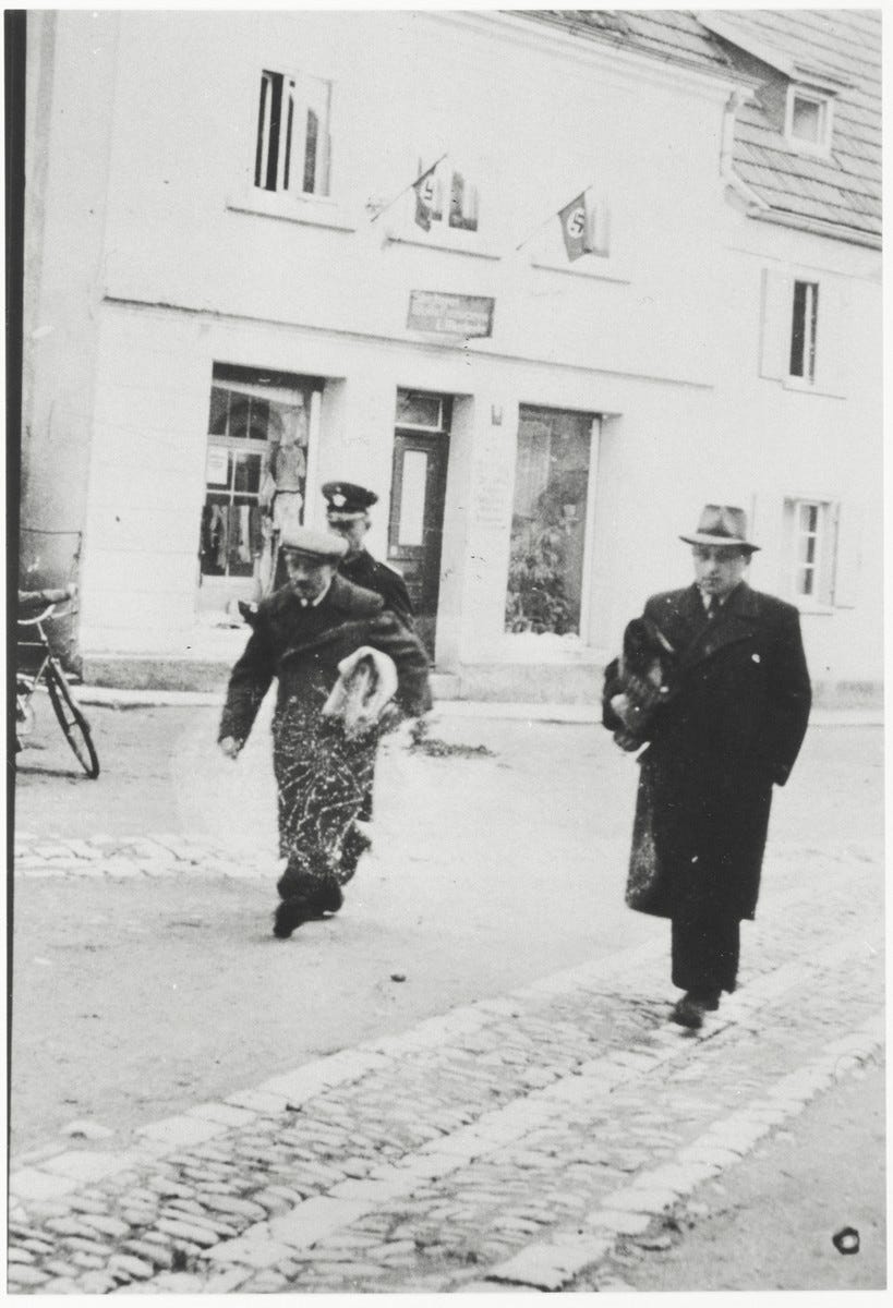Two Jewish men, carrying blanket rolls, are escorted to a police station following their arrest. Arthur Einstein is pictured on the right. On the left is Max Heimbach.