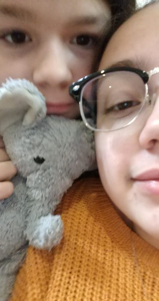 A close-up selfie of a little girl and her stuffed elephant and a woman wearing glasses and a yellow shirt. All their heads are pressed close to each other to fit into the frame of the photo.