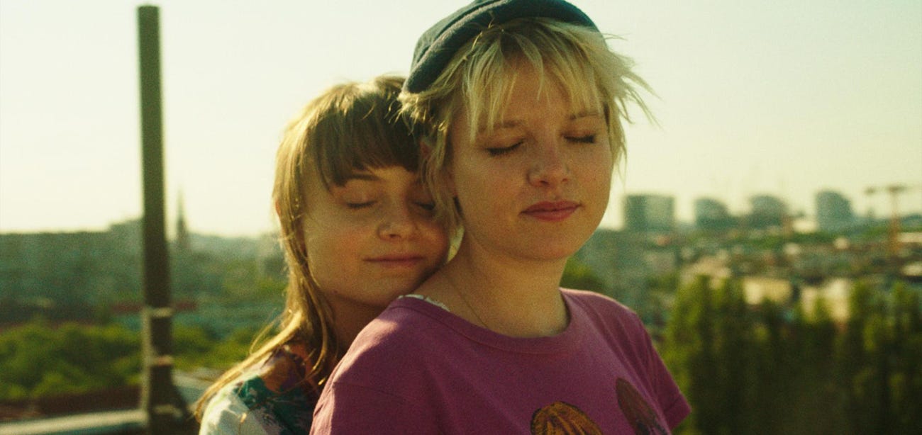 Two teenage girls with their eyes closed against a city backdrop. The shorter girl is cuddling up to the taller one.