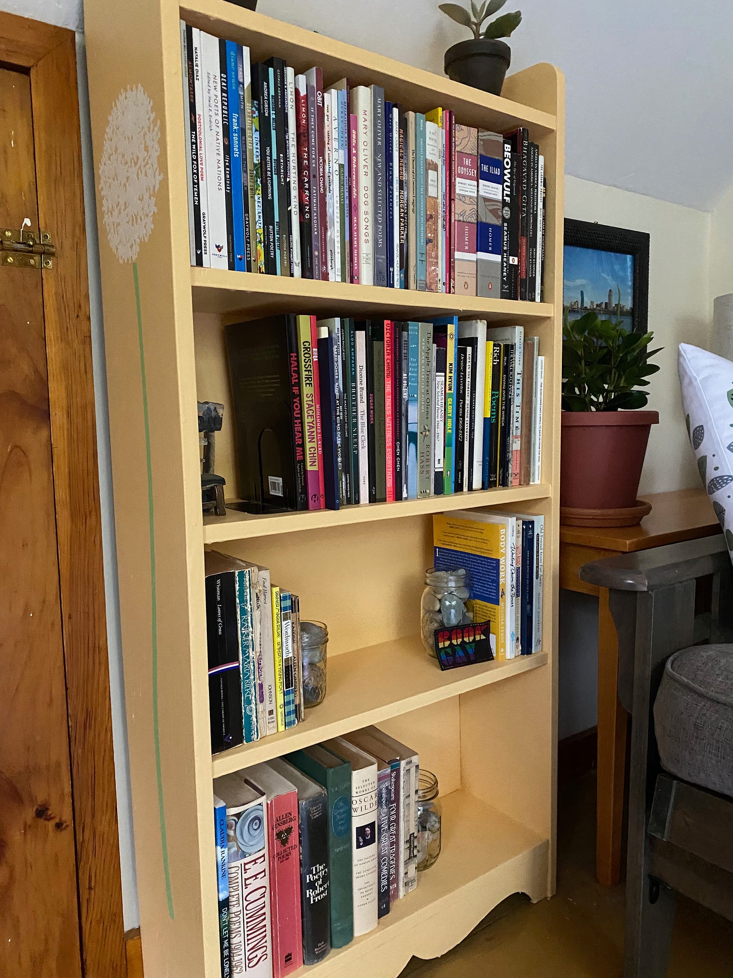 View of an orange bookcase with an allium painted on one side. It holds various shelves of poetry, rock jar bookends, and a patch that says ‘Book Gay’.