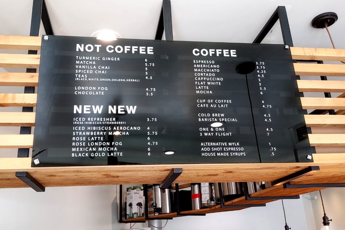 A black menu board with white block letters listing the coffee and not coffee options hangs from a pine wood shelving unit near the ceiling.