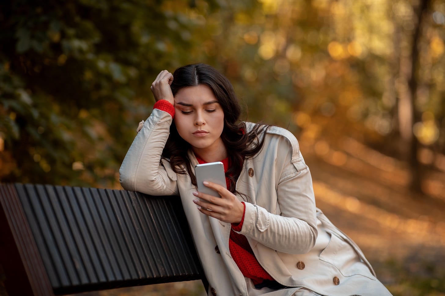 A woman sits on a bench in autumn and grimaces at her phone with a disappointed look.