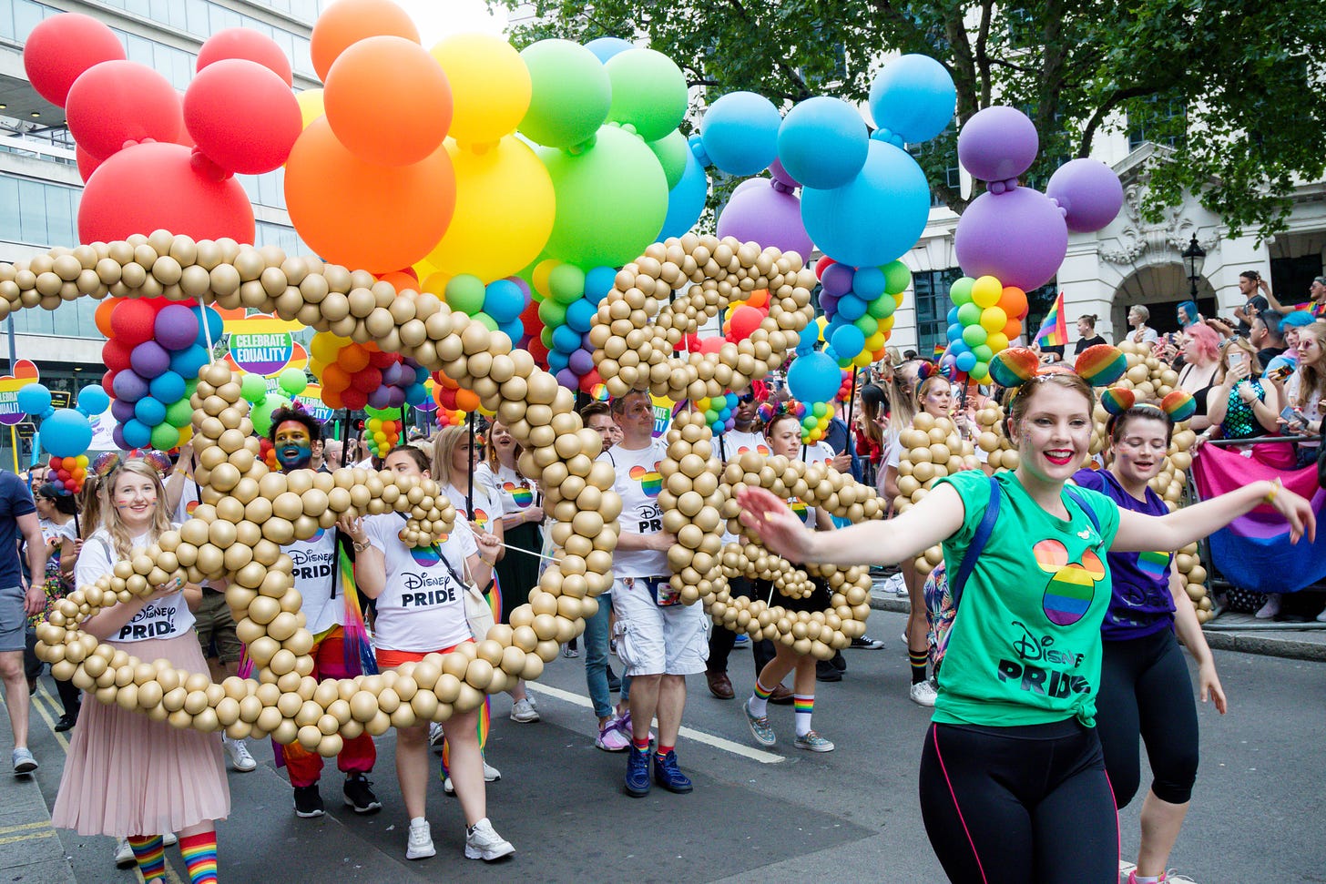 A photo of Disney fans at a pride parade. They are carrying the Disney logo spelled out in gold balloons, and a series of rainbow-colored Mickey Mouse balloons.