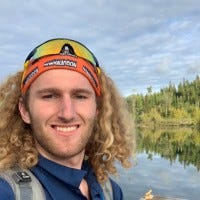 [ID: Joe Schuler, a young man with white skin and long, curly, shoulder-length blonde hair, wearing a backpack and navy blue shirt, an orange headband and ski goggles, and standing in front of a pond surrounded by trees of many shades of green and a blue sky patterned with grey and white clouds.]