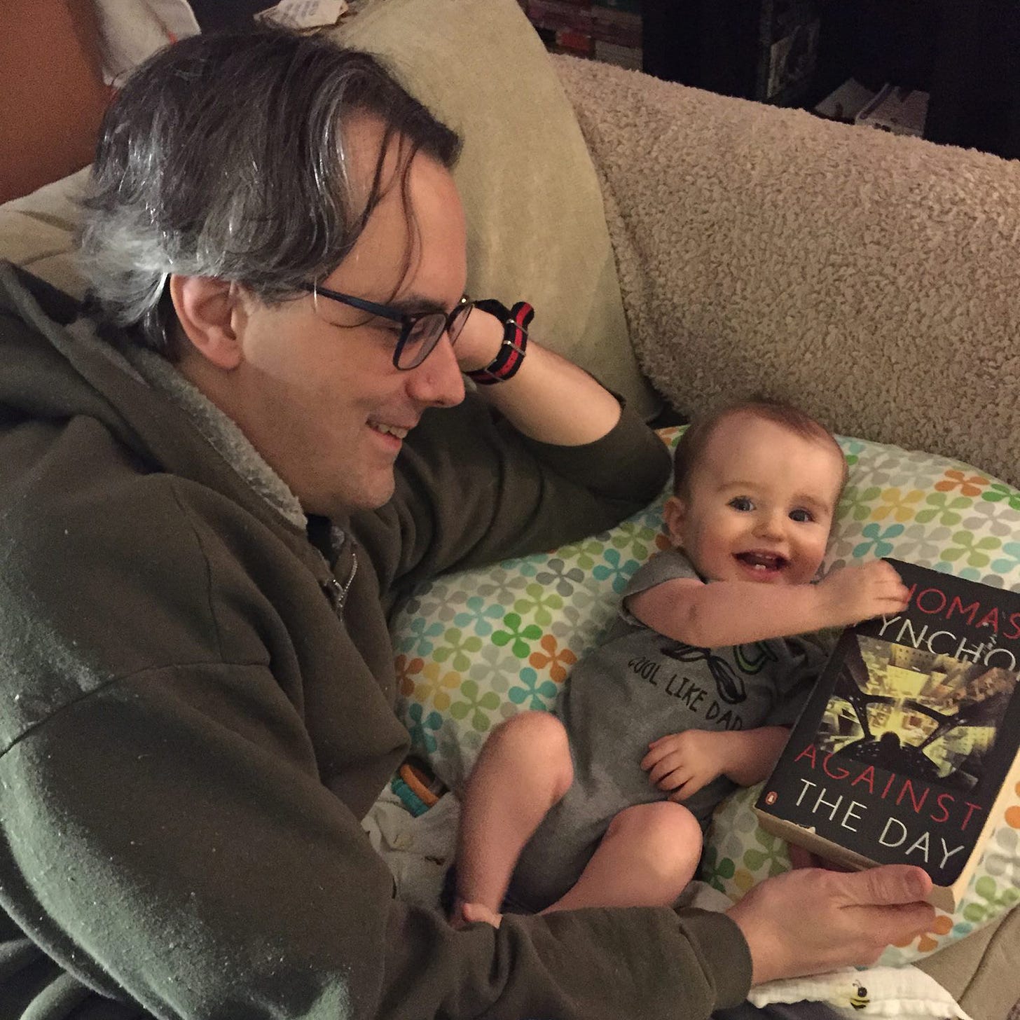 The newsletter author with his eight month old son, smiling, next to a paperback copy of Against the Day by Thomas Pynchon.